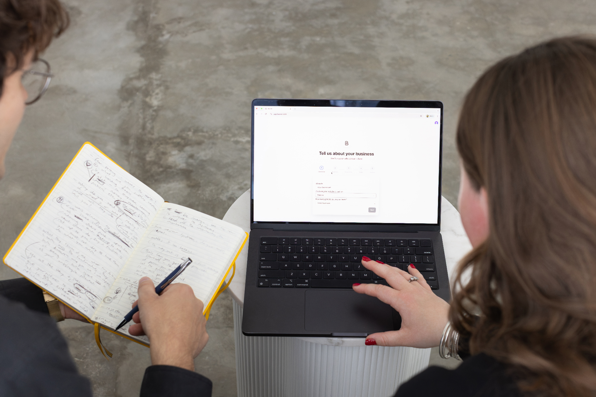 Two marketers setting up Backkr on a laptop, onboarding screen showing 'Tell us about your business' with notes and strategy planning visible